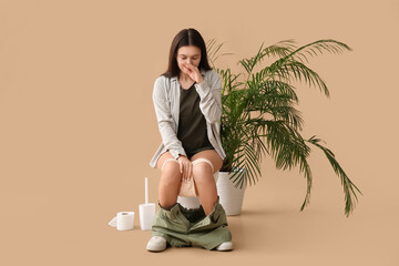 Sleepy young Asian woman sitting on toilet bowl against beige background