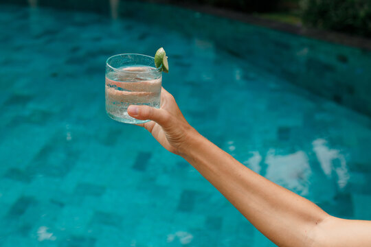 Woman holding refreshing cold drink water with ice and lime while sunbathing by the pool. Hands close up, horizontal picture