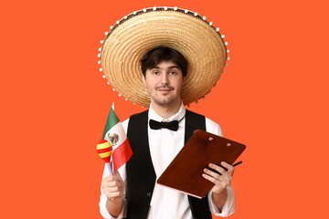Young male waiter with sombrero, flag of Mexico, maraca and clipboard on orange background