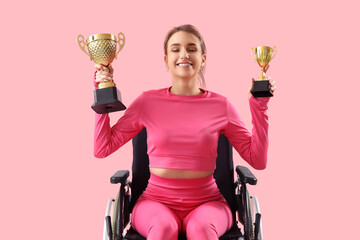 Sporty young woman in wheelchair with gold cups on pink background