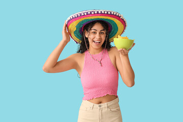 Happy young African-American woman in sombrero hat holding bowl with tasty nachos on blue background