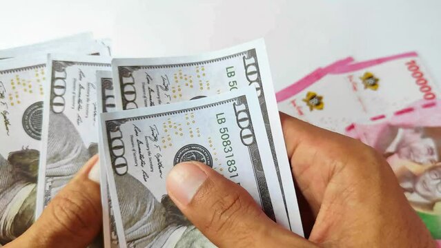 The hand of the man counting the 100 amarika dollars bill with a bunch of 100,000 rupiah bills on the table in the background.