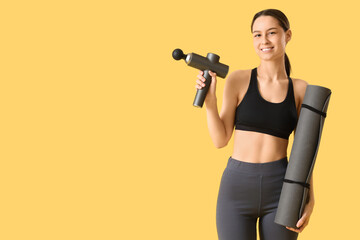 Sporty young woman with percussive massager and fitness mat on yellow background