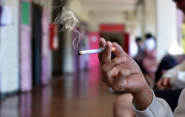 Hand holding lighted cigarette with smoking in front hallway of secondary school, concept for...