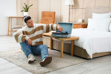 Young bearded man with headphones and record player listening to music in bedroom