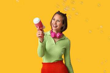 Teenage girl with soap bubble gun and headphones on yellow background