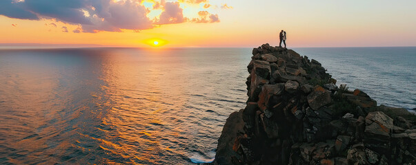 A romantic couple enjoys a breathtaking sunset from a rocky coastal peak over the vast ocean.