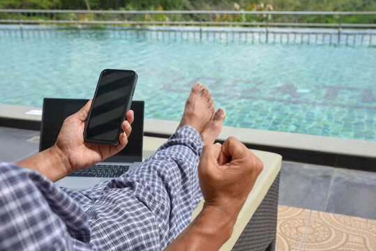 A man in a striped suit sits on a sun lounger by the swimming pool, holding a black smartphone in fists in joy. with a notebook placed - Powered by Adobe
