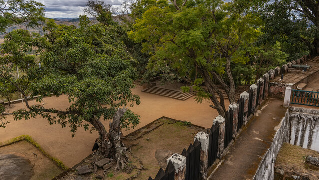 The courtyard of the palace on the Royal Hill of Ambohimanga. Green ficus trees grow around the square. In the foreground is a concrete path with a weathered wooden fence. Madagascar. Antananarivo