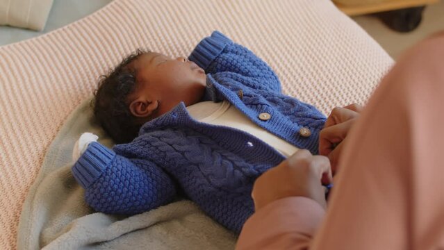 Over shoulder of unrecognizable Black woman unbuttoning blue knitted jumpsuit on tiny little baby lying on bed at home