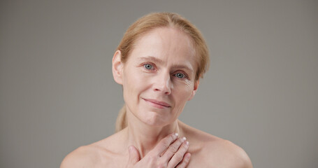 Real - people age, beauty, health and dry skin care concept - beautiful mature Caucasian middle-aged woman in her 50s, touching the skin of her face and neck, looking at the camera with a slight smile