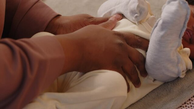 Tiny African American baby girl in cute white romper lying with her eyes closed in bed and awakening from sleep being stroked on belly by mother
