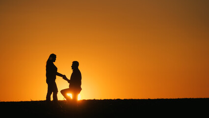 Fototapeta premium Silhouettes of a man and woman at sunset. A man is on one knee in front of a woman. Marriage proposal