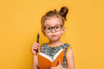 Portrait of a little girl with glasses holding a pen and notebook in her hand as she thinks, writes or takes notes on a yellow background. - Powered by Adobe