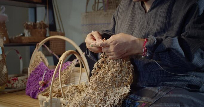 Slow motion scene while an Asian female artisan who is a local villager knitting folk basket made from leftover paper band to sell as extra income in the basketry shop shop.