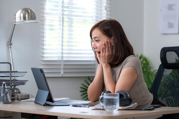 A woman is sitting at a desk with a tablet. She is looking at the laptop and she is surprised