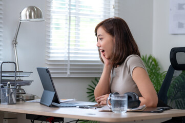 A woman is sitting at a desk with a tablet. She is looking at the laptop and she is surprised