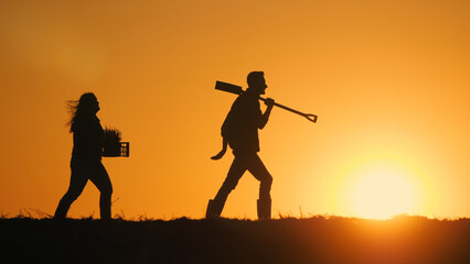 A couple of farmers with equipment walks through a field at sunset.