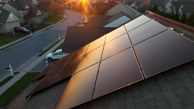 Solar panels reflecting golden hour sunlight on shingle roof of modern home with renewable energy source. Aerial tilt up revealing sunburst during sunset.