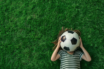 Overhead view of a child laying on green grass holding a soccer football ball