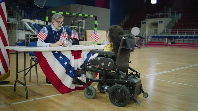 Woman with spinal muscular atrophy in electric wheelchair consults with worker in polling station. Political races of US presidential candidates. National Election Day in United States. Dolly shot.