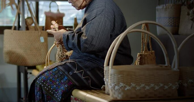 Slow motion scene while an Asian female artisan who is a local villager knitting folk basket made from leftover paper band to sell as extra income in the basketry shop shop.