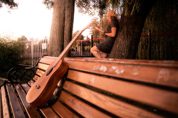 Woman reading a book in a park during the evening, with a guitar on the bench