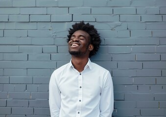 Obraz premium Joyful Young African American Man Leaning Against Blue Brick Wall in White Shirt