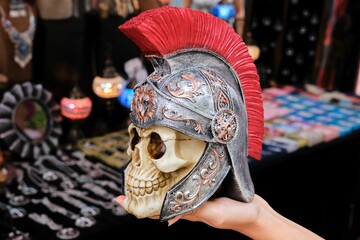 Hand is displayin a skull with Roman gladiator helmet against a backdrop of various objects