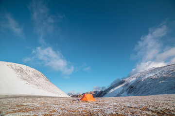 Orange tent on snow-covered stony pass in sunlight. Snowy stone hills and rocky mountain ridge at early morning. Red tent in high mountains in freshly fallen snow. Low clouds in blue sky at sunrise.
