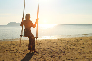 Young woman sits and swings on a rope swing on the seashore and looks at the sunset and horizon....