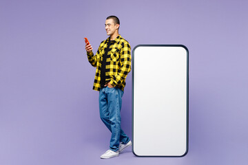 Full body side profile view young middle eastern man wears yellow shirt casual clothes big huge blank screen mobile cell phone with workspace area use smartphone isolated on plain purple background.
