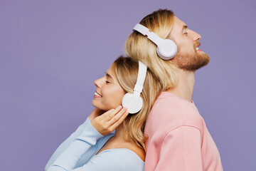 Side view close up young couple two friends family man woman wear pink blue casual clothes together listen to music in headphones stand back to back isolated on pastel plain purple background studio.