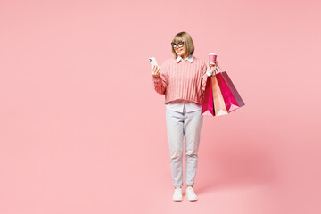 Full body elderly young woman wear casual clothes hold in hand paper package bags after shopping use mobile cell phone isolated on plain light pastel pink background Black Friday sale buy day concept