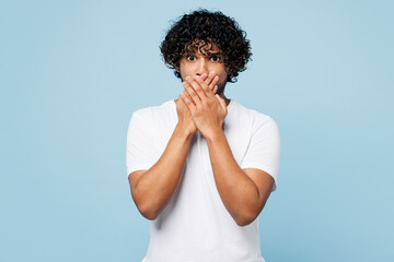 Young shocked surprised astonished sad Indian man he wears white t-shirt casual clothes cover mouth with hands isolated on plain pastel light blue cyan background studio portrait. Lifestyle concept.
