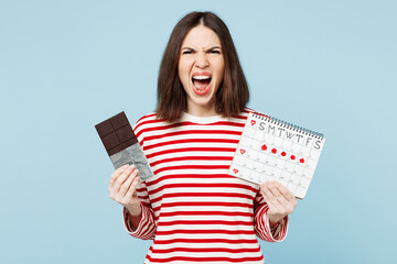 Young angry mad woman she wears red casual clothes eat sweet chocolate bar hold female periods pms calendar checking menstruation days isolated on plain blue background. Medical gynecological concept. - Powered by Adobe