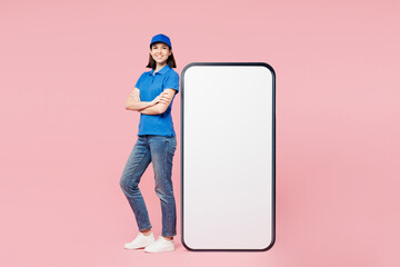 Full body delivery employee woman wears blue cap t-shirt uniform workwear work as dealer courier stand near big huge blank screen mobile cell phone with empty area isolated on plain pink background.