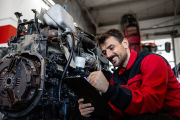 Mechanic holding checklist and repairing tractor engine in workshop.