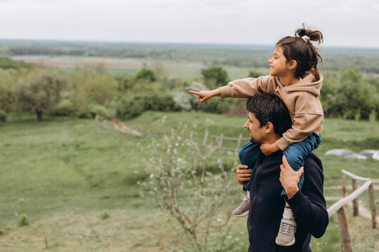 a child and his father are having fun in nature, a girl with dark hair in a sweatshirt sits on her dad&rsquo;s shoulders and laughs