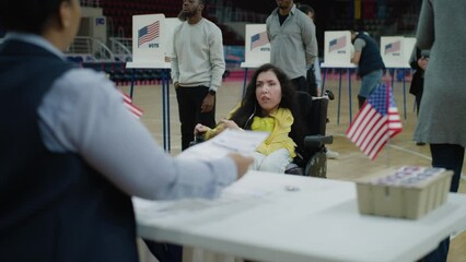Woman with SMA in motorized wheelchair comes and talks with African American polling officer. Political races of US presidential candidates in polling station. National Election Day in United States.
