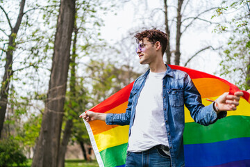 A smiling gay man casually holds the colorful LGBT flag, surrounded by serene park scenery.