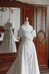 A white dress is displayed in a room with a wooden dresser. The dress is hanging on a mannequin and...