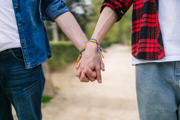 Close-up view of a male couple's hands interlocked, adorned with vibrant LGBT bracelets in daylight.