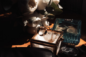 A couple of gold rings are displayed in a glass box on a table. The rings are surrounded by a bouquet of white flowers and a bottle of perfume. Concept of romance and celebration