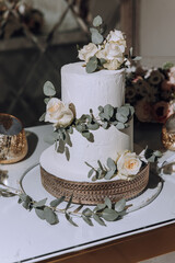 A white cake with a green leaf and white roses on top. The cake is placed on a table with a gold bowl and a vase