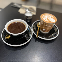 A glass of latte and a cup of coffee on a table with blurry background