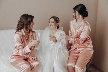 Three women in pink pajamas are sitting on a bed and drinking wine. They are smiling and seem to be...