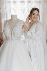 A woman is standing in front of a wedding dress, getting ready to put it on. The dress is white and has a lace design. The woman is smiling and seems excited about the upcoming wedding