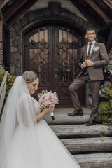 A bride and groom stand in front of a large wooden door. The bride is holding a bouquet of flowers, and the groom is wearing a suit. Scene is romantic and intimate