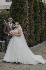A bride and groom are posing for a picture in front of a bush. The bride is wearing a white dress and the groom is wearing a suit. The couple is holding a bouquet and a bouquet of flowers